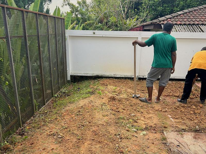 Two workers, seen from behind, clearing and levelling bare orange soil beside the mesh fence at the start of the job.